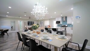 A modern dining area with a chandelier, a kitchen in the background, and a notice sign on the wall.