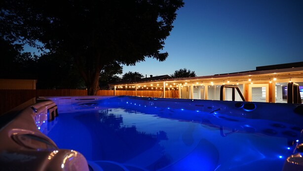 A hot tub with blue lighting, surrounded by a wooden deck and a building with string lights.