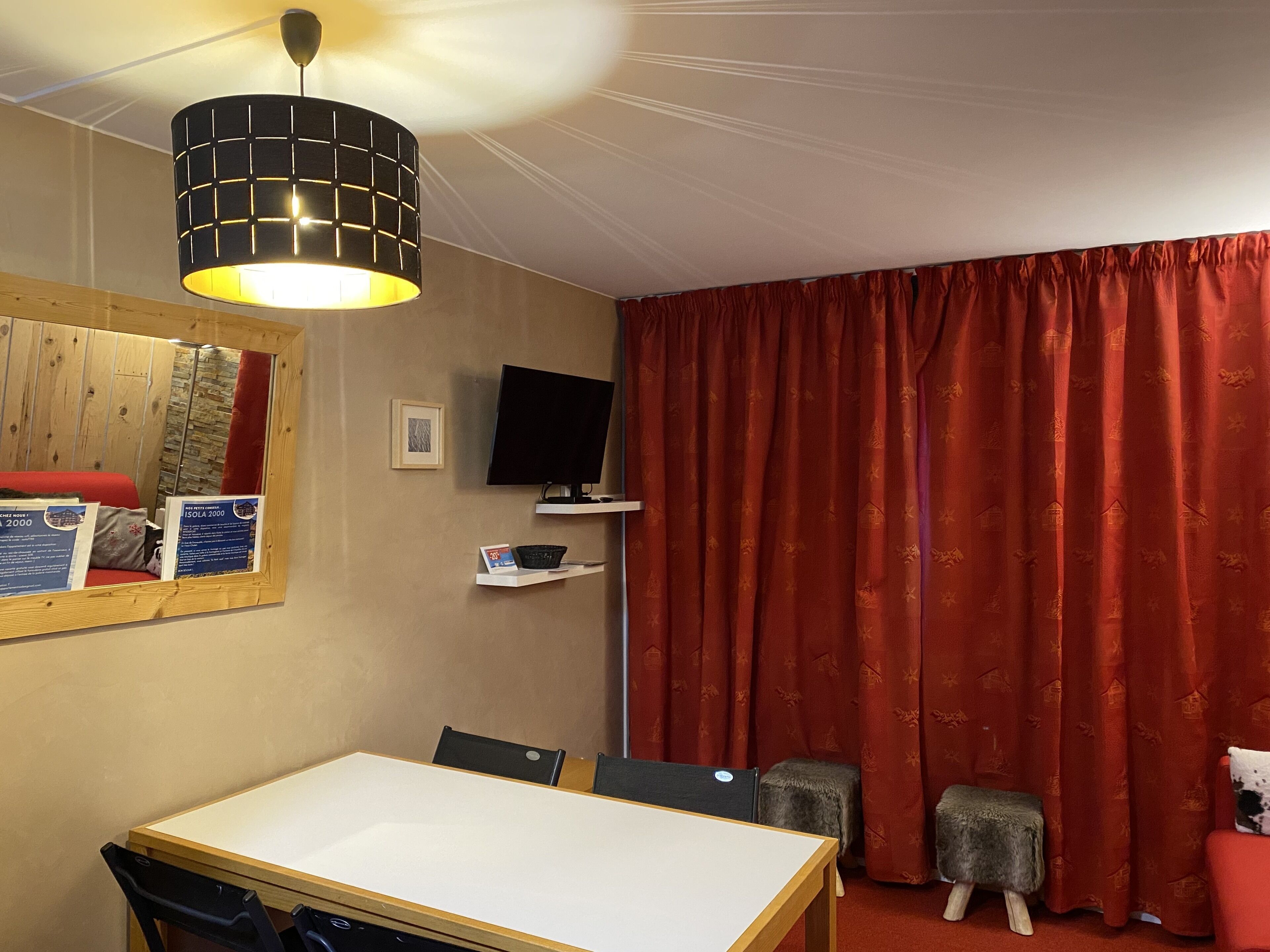 A hotel room with a wooden ceiling, a television on a shelf, a table with chairs, a red curtain, and a mirror on the wall.