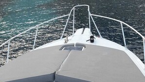 A boat's bow with a grey deck and metal railings, heading towards a rocky coastline with white buildings with blue windows. - Egadi Islands Boat Excursions (Trapani)