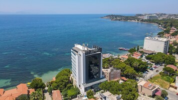 Un hotel moderno con un amplio ventanal de vidrio, situado cerca de una zona costera con aguas azules cristalinas y barcos.