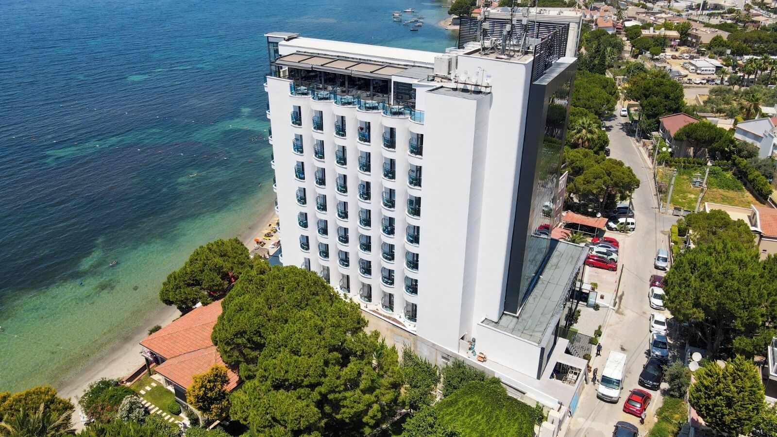A high-rise hotel with a rooftop terrace, adjacent to a beach and a parking area.