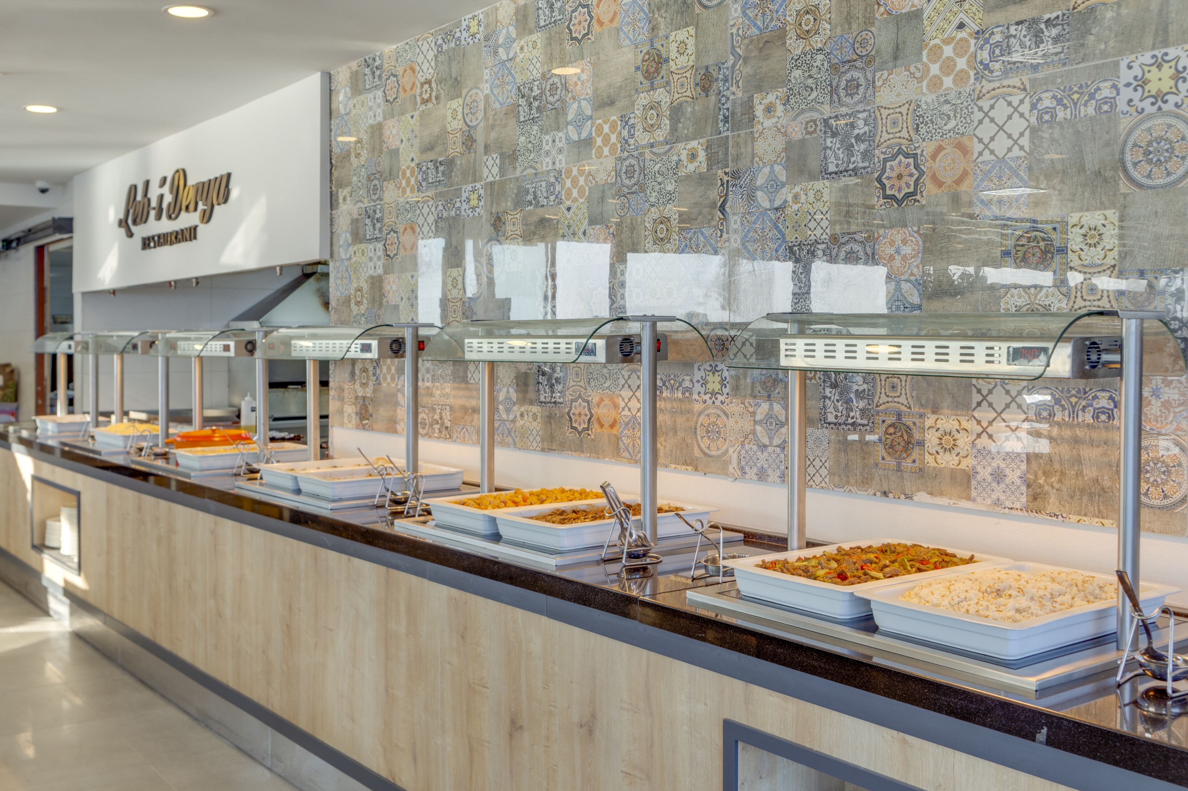 A buffet counter with various food items and a tiled wall with a restaurant sign in the background.