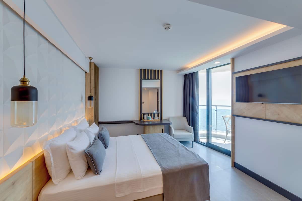 Ein modernes Hotelzimmer mit einem großen Bett, einem Schreibtisch mit Spiegel und Meerblick durch große Fenster.