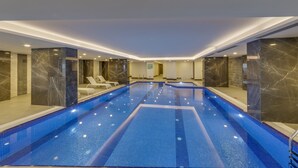Une vaste piscine intérieure, dont le sol et les murs sont recouverts de carreaux, entourée de piliers de marbre et de chaises longues.