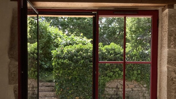 An open door leading to a patio with stone flooring, a white metal table and chairs, and a stone wall with greenery.
