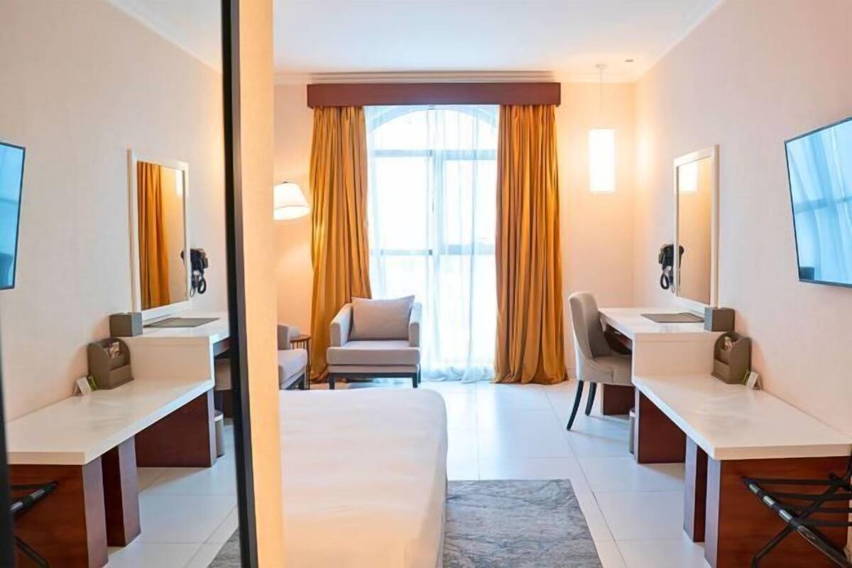 A hotel room with a bed, two desks, a chair, a mirror, and a window with curtains.