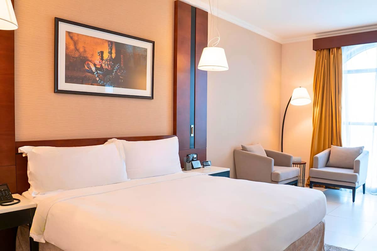 A hotel room with a large bed, a desk with a phone, a chair, a lamp, and a painting on the wall.