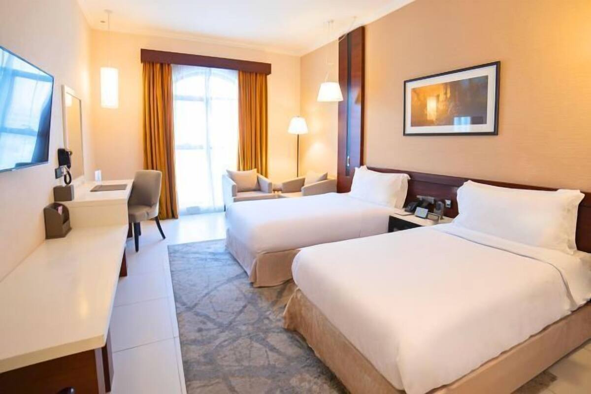 A hotel room with two beds, a desk, a chair, a TV, and a lamp.