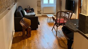 Un ambiente compatto con divano, un tavolino, una sedia e una televisione. Una finestra offre luce naturale, e una parete in pietra si trova su un lato.