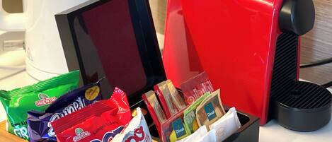 Une machine à café, une cafetière blanche et une caisse en bois contenant des paquets de collations.