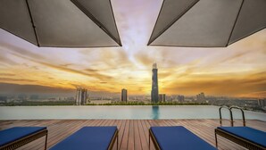 Una piscina a sfioro con terrazza in legno e poltrone relax, con vista sullo skyline di una città al tramonto.
