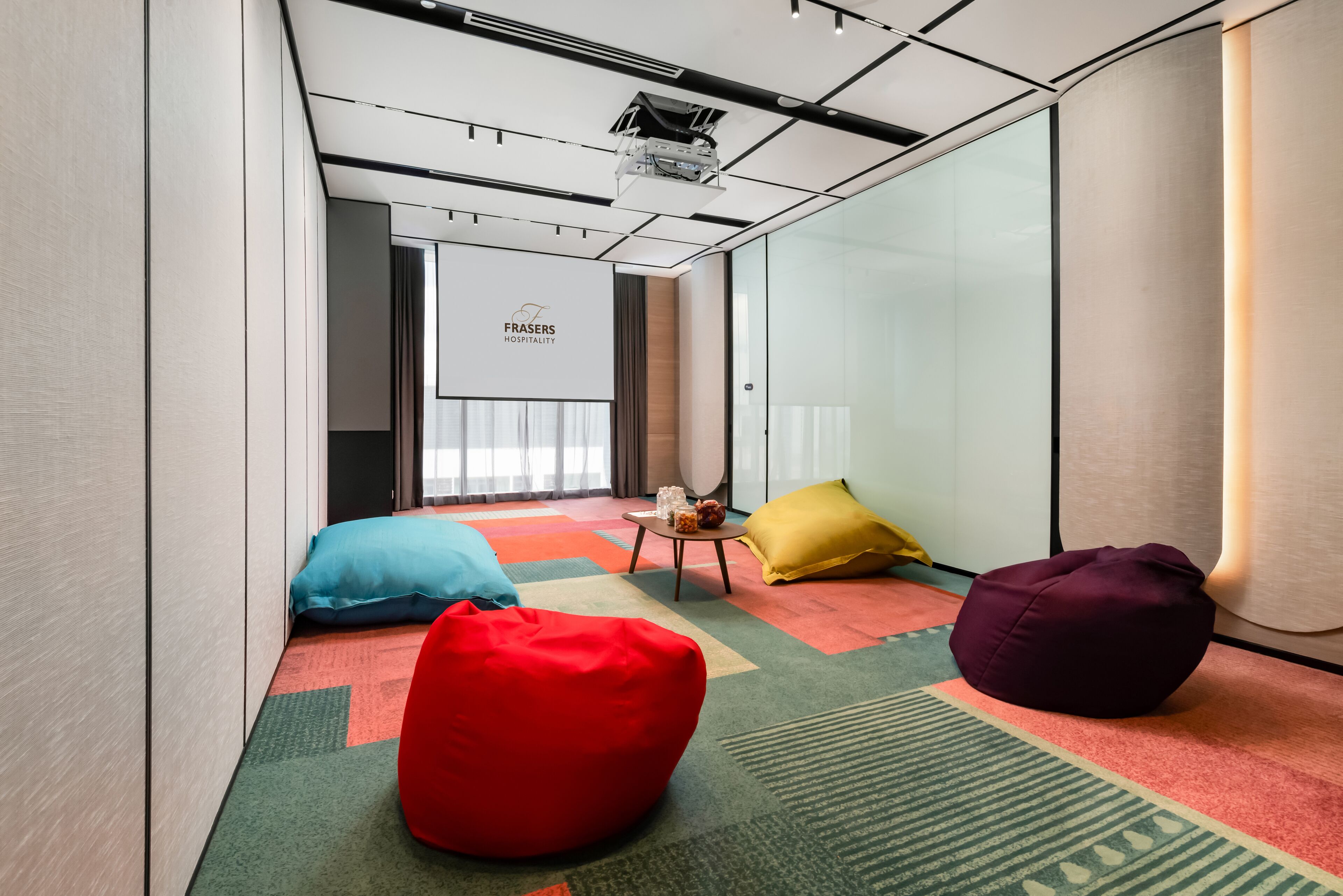 Una habitación de hotel moderna con una pantalla de proyector, coloridos pufs y piso alfombrado.