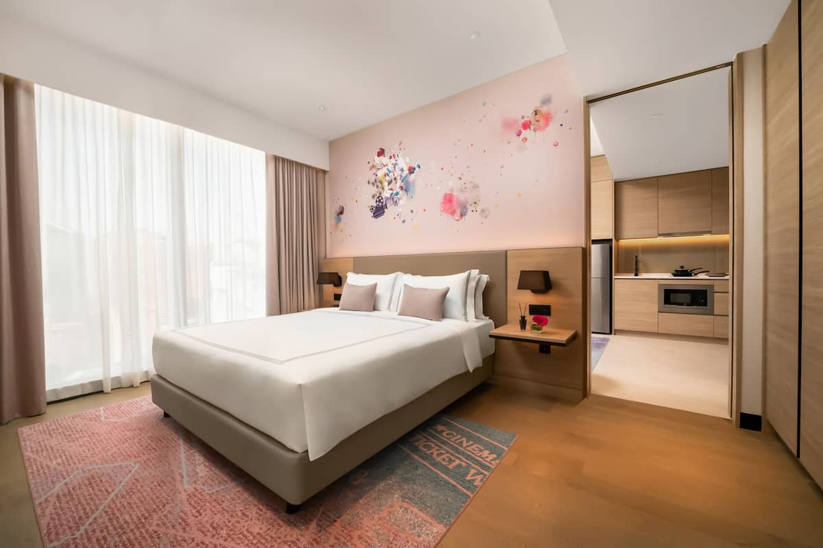 A modern hotel room with a large bed, a small bedside table, a decorative wall, and a view of the kitchen.