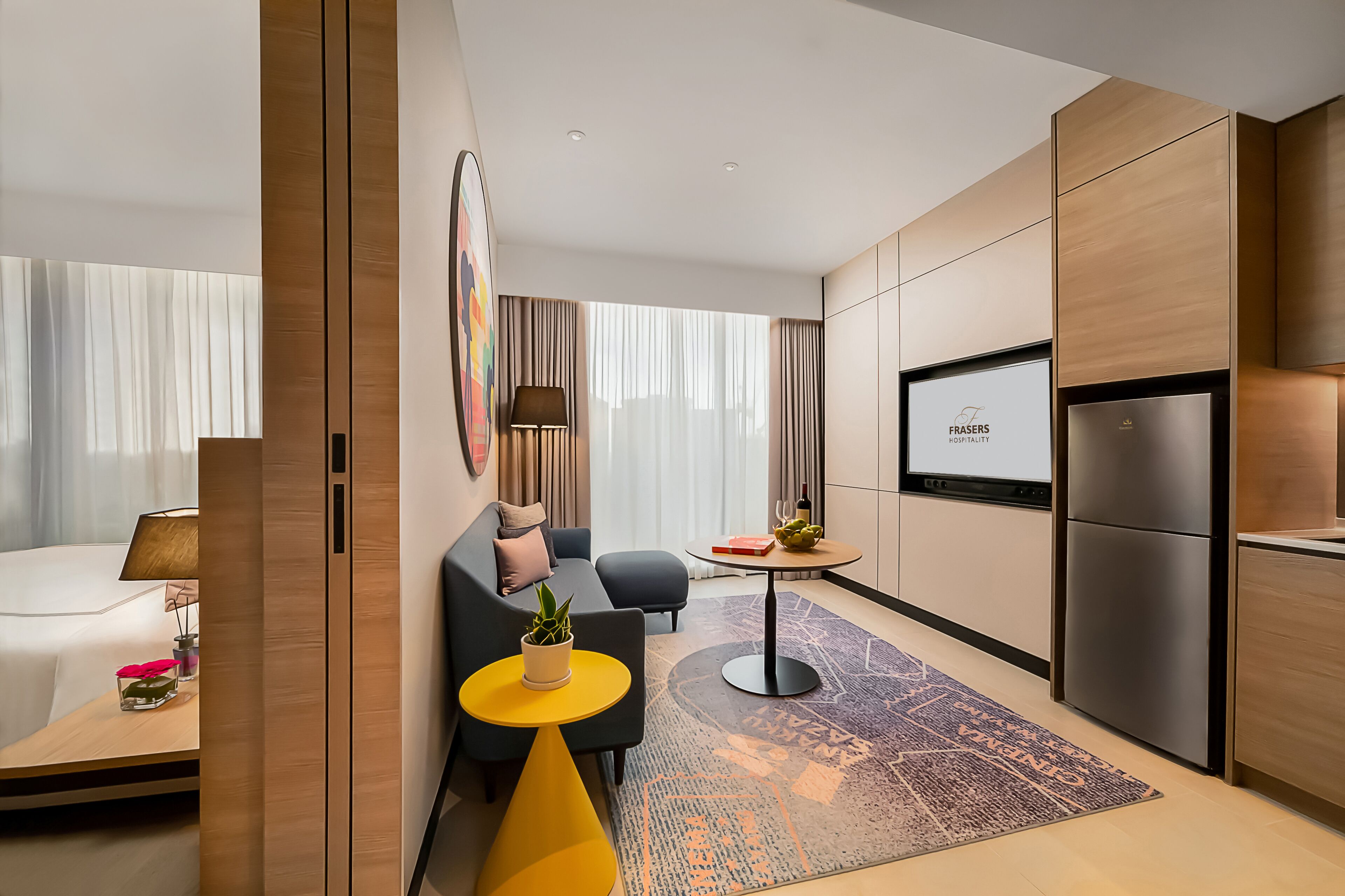 Una habitación de hotel moderna con cama, sofá, una mesita con lámpara, televisor y refrigerador.