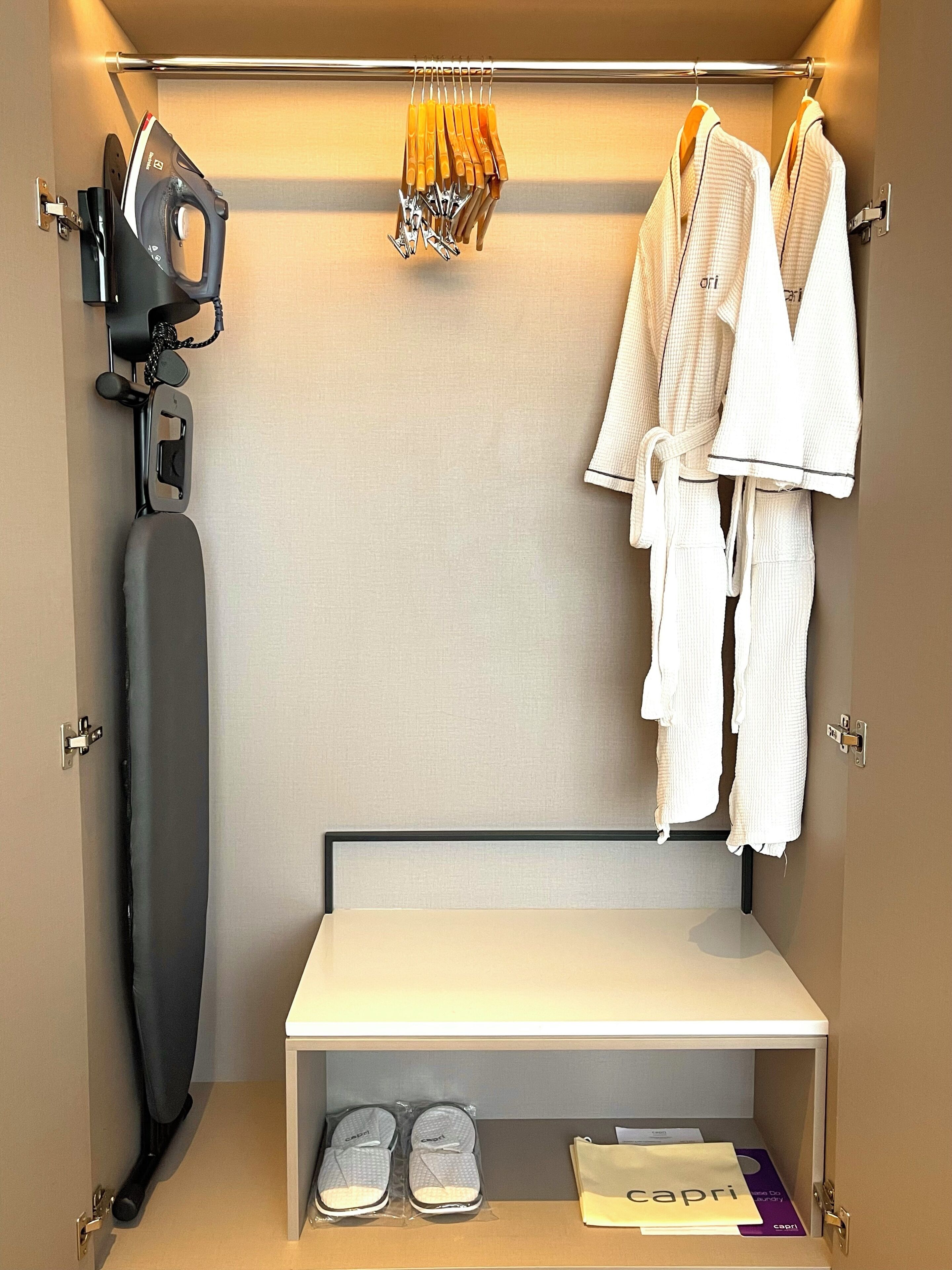 A hotel room with a built-in ironing station, a rack of robes, a pair of slippers, and a small table.