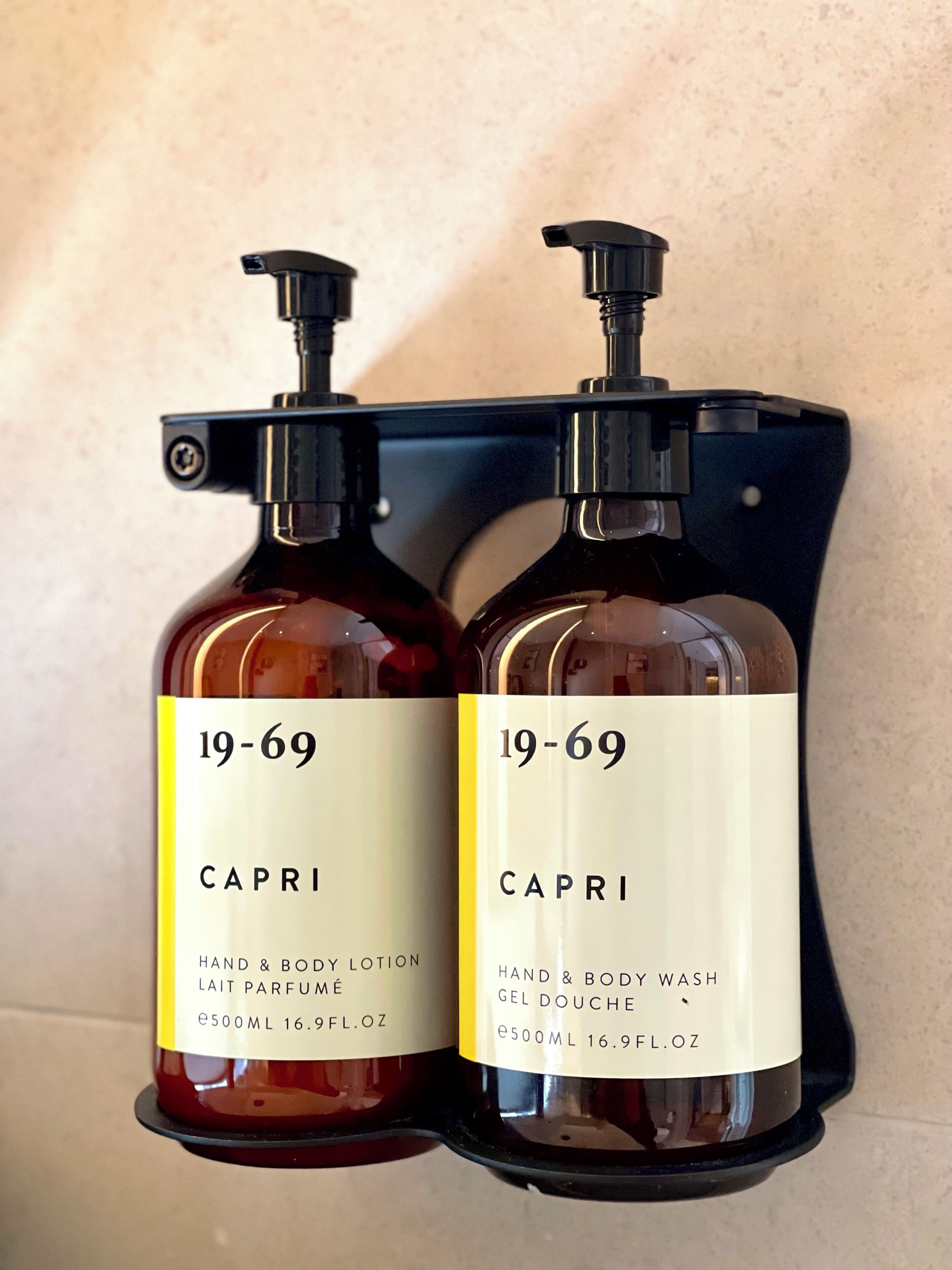 Two bottles of Capri hand and body lotion and wash gel, each with a pump dispenser, mounted on a black holder against a textured wall.
