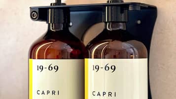 Two bottles of Capri hand and body lotion and wash gel, each with a pump dispenser, mounted on a black holder against a textured wall.