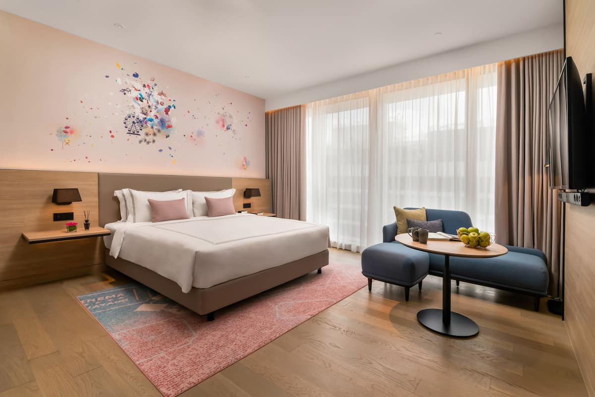 A modern hotel room with a large bed, a small sofa, a round table with fruit, and a wall art piece.