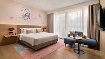 Una habitación de hotel moderna con una cama grande, un pequeño sofá, una mesa redonda con fruta y una obra de arte en la pared.