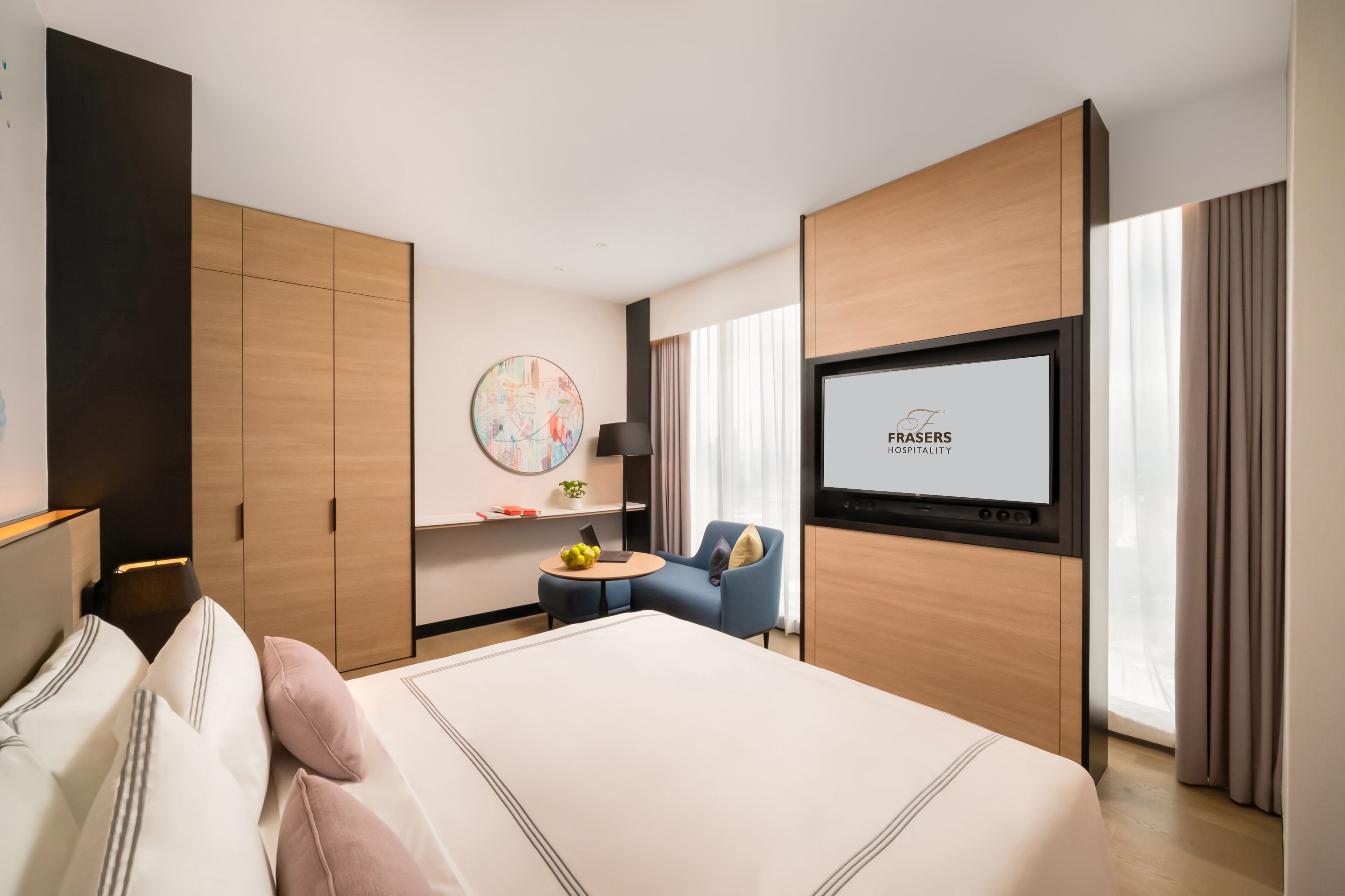 Une chambre d’hôtel avec un grand lit, un écran de télévision affichant le logo « Frazers Hospitality », une petite table ronde avec un ordinateur portable et des fruits, et une chaise bleue.