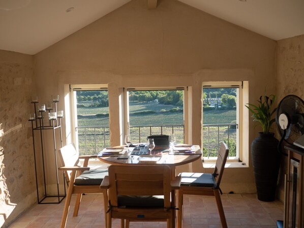 Un comedor con mesa y sillas, un ventilador y vistas a viñedos a través de amplios ventanales.