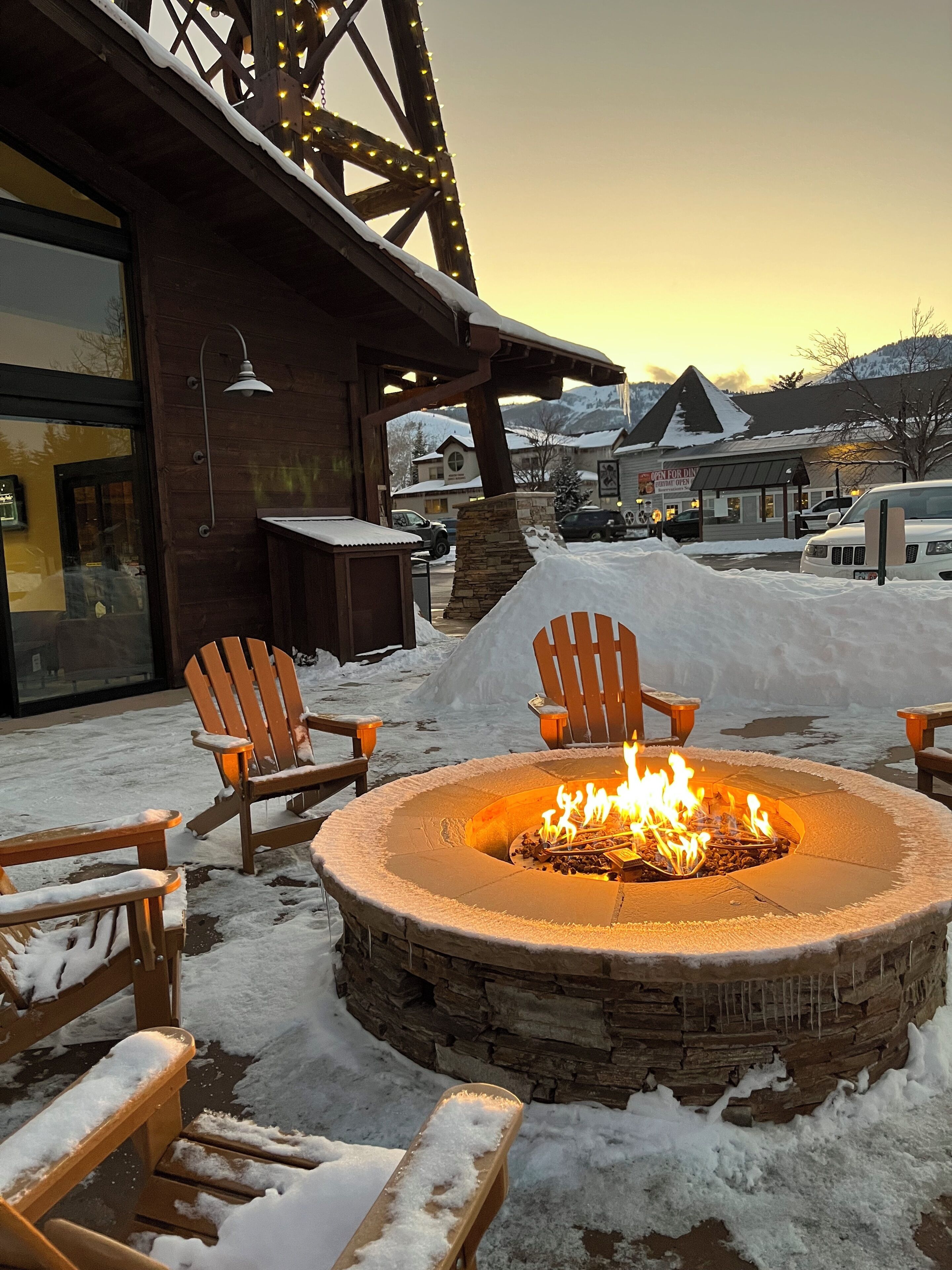 A stone fire pit with chairs around it, set in a snowy outdoor area with a building in the background.