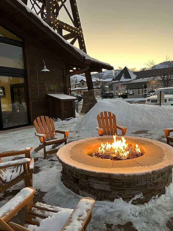 A stone fire pit with chairs around it, set in a snowy outdoor area with a building in the background.