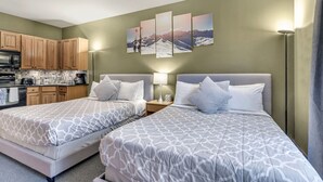 A hotel room with two beds, a kitchenette, and a painting of a mountain landscape on the wall.