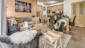 Una sala de estar acogedora con un sofá, una mesa de centro y una manta de piel sintética. Hay una zona de comedor con una mesa y sillas, y una cocina al fondo.