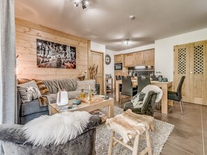 A cozy living room with a sofa, a coffee table, and a fur throw. There's a dining area with a table and chairs, and a kitchen in the background.