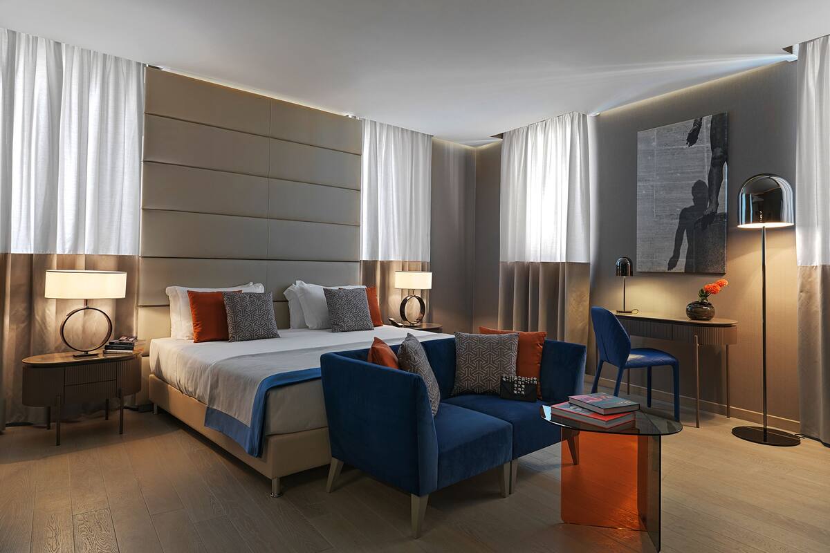 A modern hotel room with a large bed, a sofa, a desk, and a chair. There are two lamps and a painting on the wall.