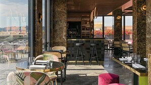 A modern bar area with a view of the city, featuring a bar counter, stools, and a seating area with tables and chairs. - Orazio Palace Hotel (Rome)
