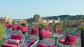 Eine Dachterrasse mit Korbstühlen und Tischen, mit Blick auf eine Stadtlandschaft mit historischen Gebäuden und Grünflächen.