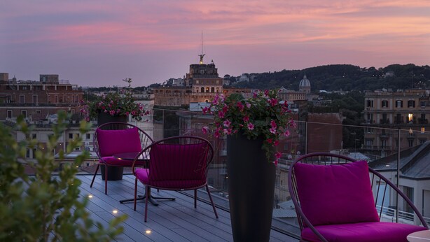 Terraza en la azotea con dos sillas rosas y una maceta grande con flores, con vista a un paisaje urbano de edificios históricos.