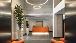 Un área de recepción moderna con un mostrador central naranja, dos plantas en macetas y un par de sillones anaranjados.