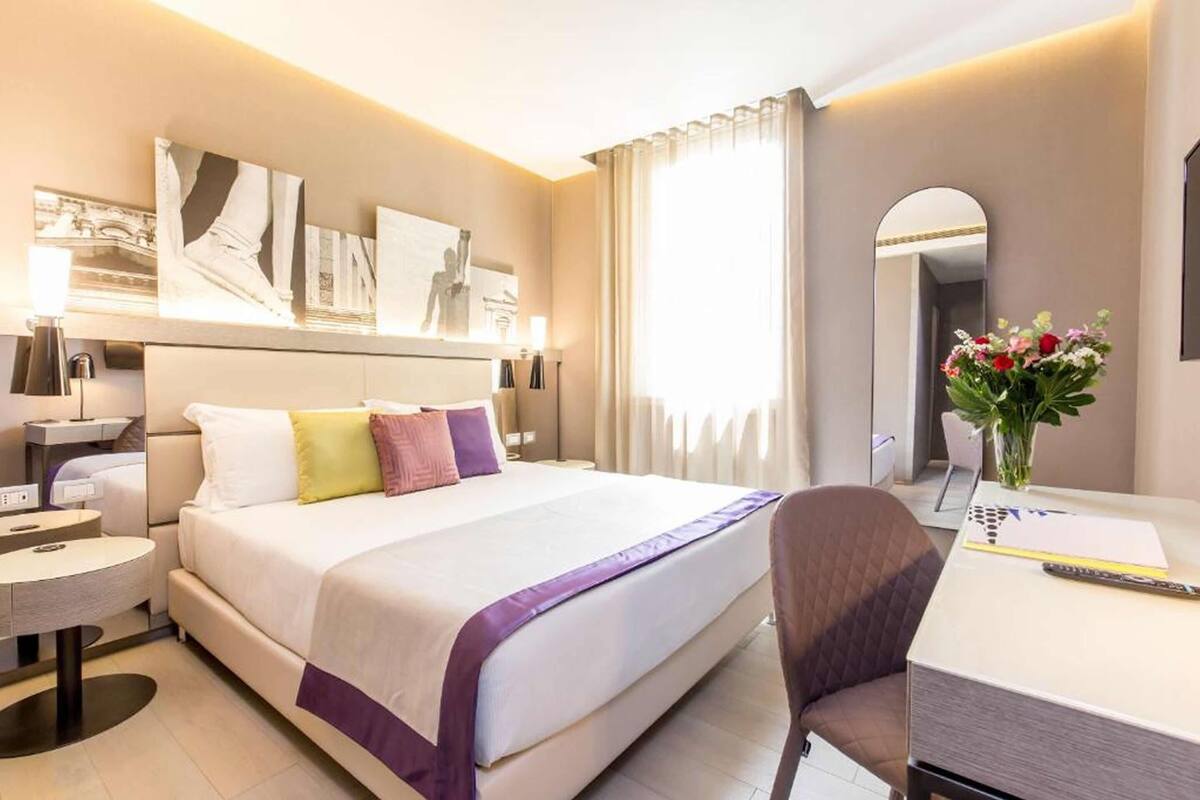 A modern hotel room with a large bed, a desk, and a chair. There is a mirror on the wall and a vase with flowers on the desk.