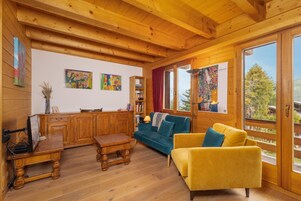 Interior de cabana de madeira com escrivaninha de madeira, um sofá azul, uma poltrona amarela e uma estante de livros.