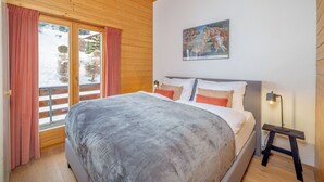 A bedroom with a large bed, wooden ceiling, a painting on the wall, and a view through a window. - Chalet Mouettes (Riddes)
