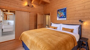 Un dormitorio con paneles de madera, una cama, una mesita de noche, un cuadro colgado en la pared y un baño visible a través de una puerta abierta.