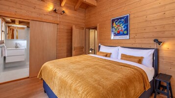 Quarto com painéis de madeira, uma cama, uma mesa de cabeceira, um quadro pendurado na parede e um banheiro visível por uma porta aberta.