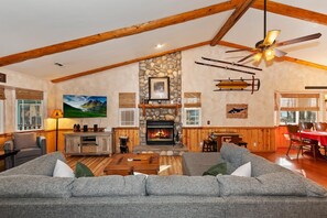 A spacious living room with a fireplace, a large sofa, wooden beams, and a ceiling fan.