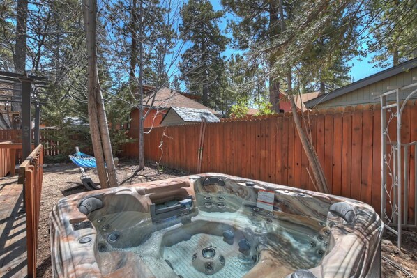 Un jacuzzi en un patio trasero con una valla de madera, árboles y una casa al fondo.