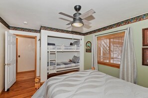 A bedroom with a bunk bed, a ceiling fan, a window with curtains, and a door leading to another room.