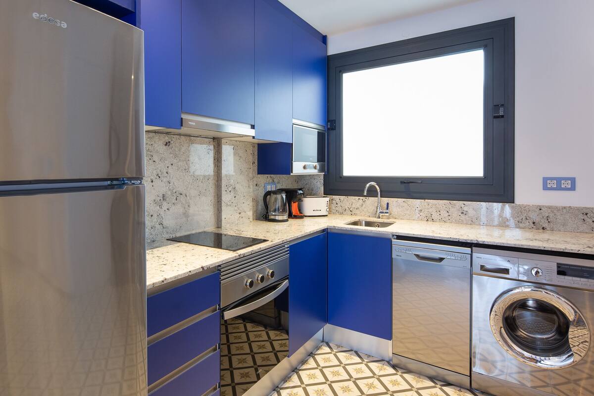 Una cocina moderna con armarios azules, electrodomésticos de acero inoxidable, una ventana y un salpicadero de azulejos.