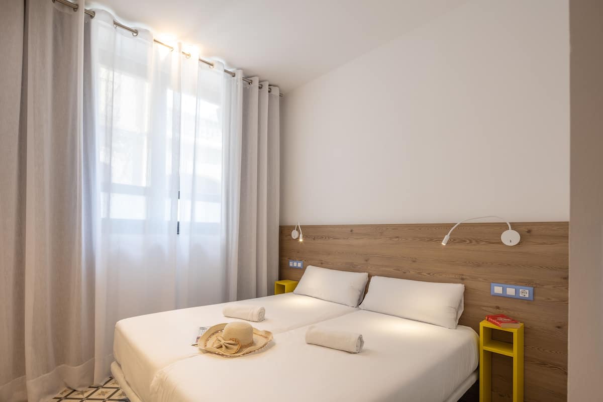 Habitación de hotel con cabecera de madera, cama con sábanas blancas, mesita de noche amarilla y ventana con cortinas translúcidas.