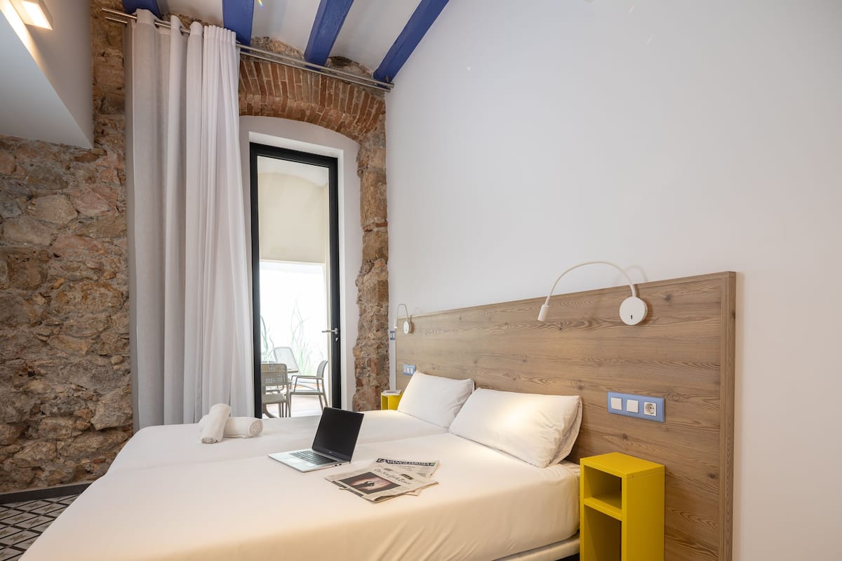 Un dormitorio con pared de piedra, cabecera de madera, cama con sábanas blancas, una computadora portátil y una mesita de noche amarilla.