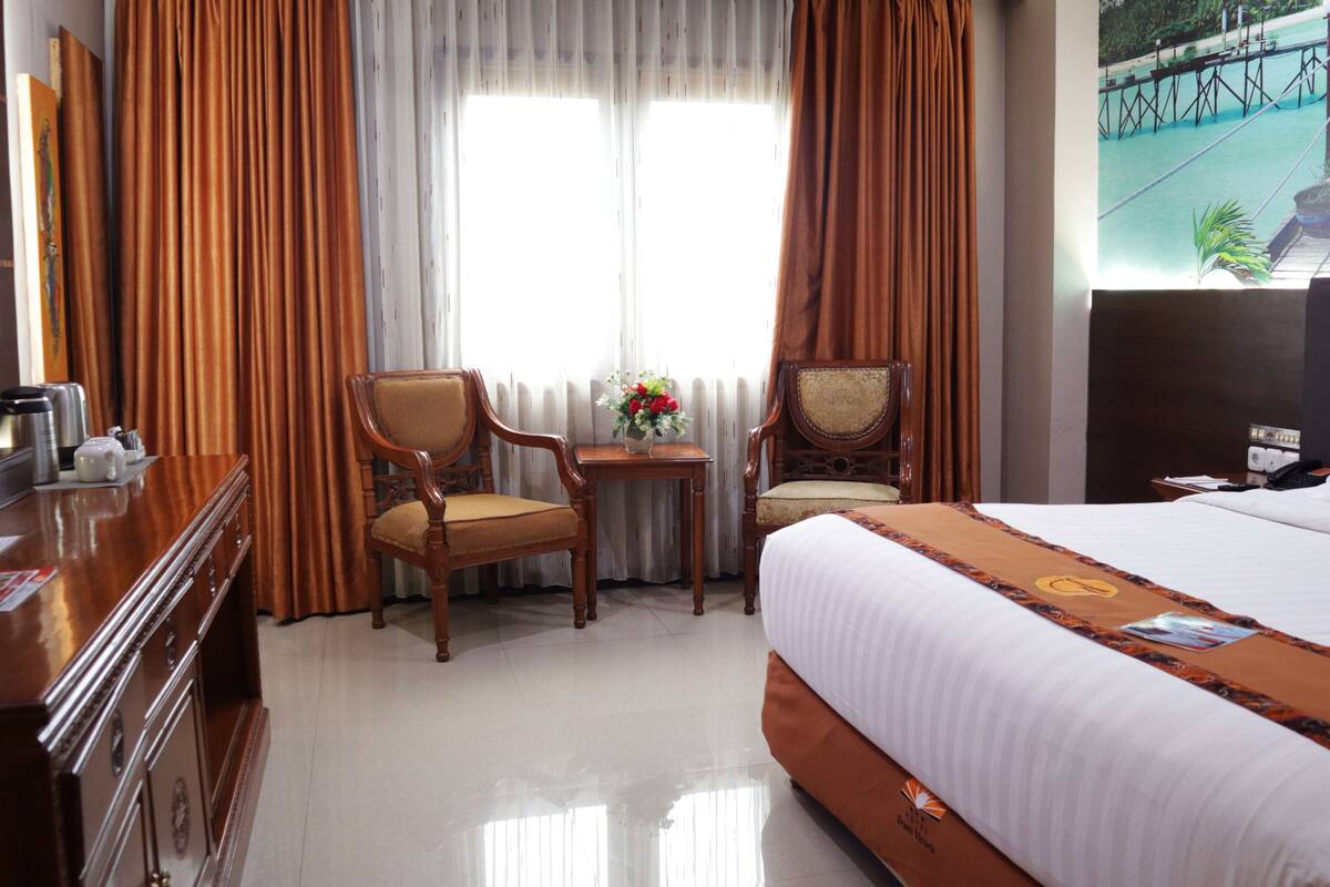 A hotel room with a bed, two chairs, a small table with a vase of flowers, a desk with a telephone, and a TV on the wall.