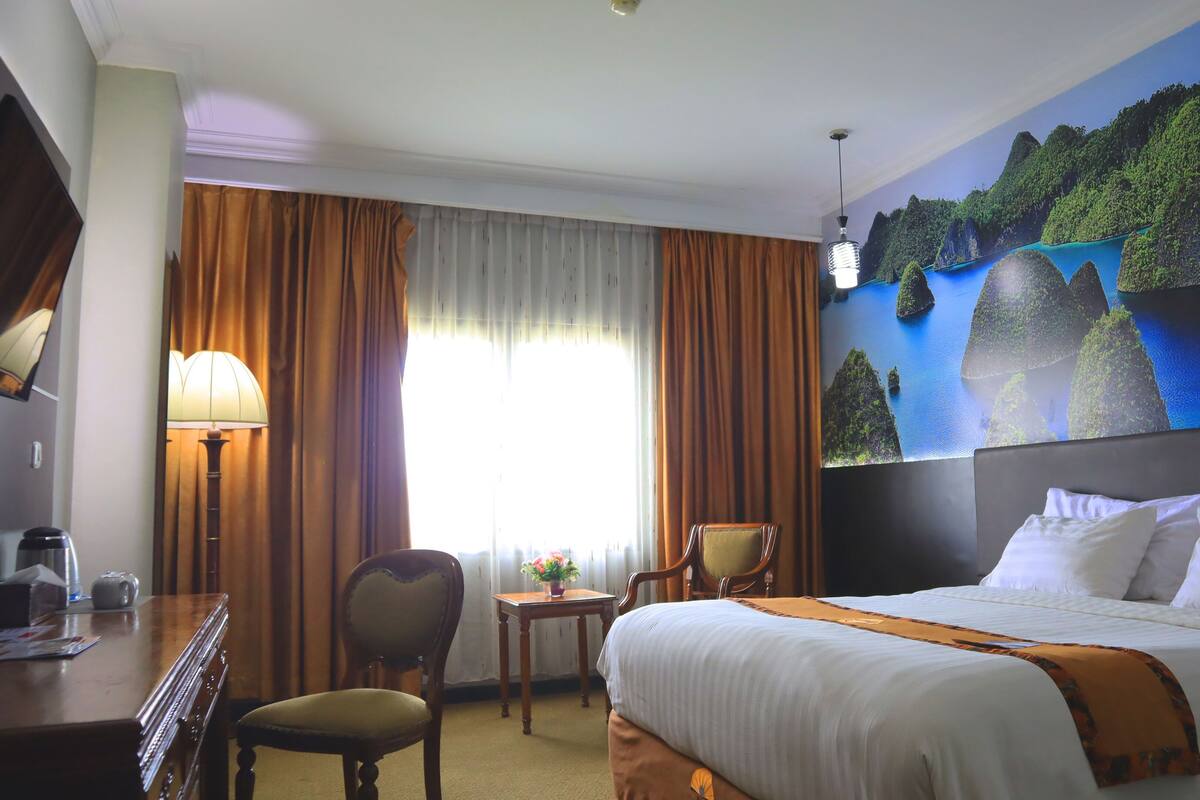 A hotel room with a bed, a desk, a chair, a lamp, and a painting of a landscape.