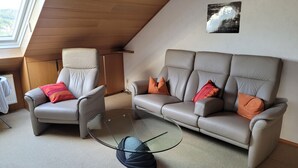 A modern living room with a high ceiling, a glass coffee table, a grey sofa, and a leather armchair. - Gepflegte Dachgeschosswohnung im 2-familienhaus in Ruhiger Wohnlage (Bad Kissingen)